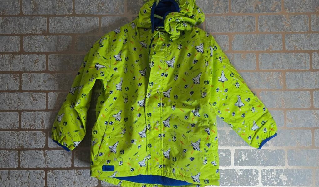 A bright green children’s winter jacket with blue and white sailboat and fish patterns hangs on a hook against a light-colored brick wall. The jacket has a hood, blue inner lining, and a front zipper with a flap covering it.