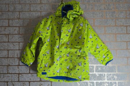 A bright green children’s winter jacket with blue and white sailboat and fish patterns hangs on a hook against a light-colored brick wall. The jacket has a hood, blue inner lining, and a front zipper with a flap covering it.