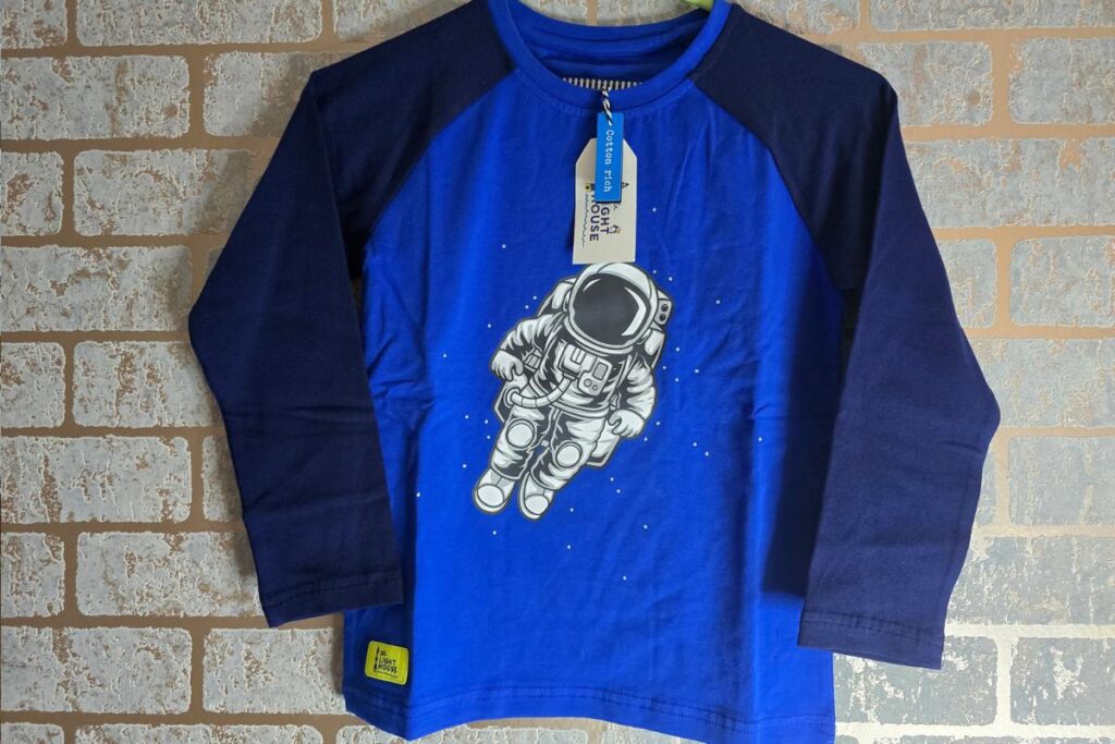 A blue long-sleeve children’s shirt with dark blue sleeves hangs on a brick wall. The front features a graphic of a white astronaut floating in space. A tag is attached to the collar, and a yellow patch is sewn near the lower left hem.