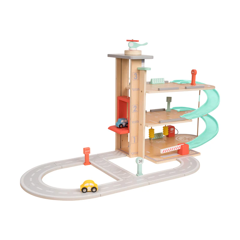 A wooden toy parking garage with three levels, a spiral ramp, a red elevator, a green helicopter on the roof, road signs, and three small vehicles (yellow, blue, red) driving on a gray and white road track that connects to the garage.