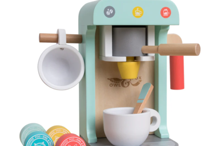 A wooden toy coffee machine in pastel colors with a cup, spoon, and wooden coffee pods. The machine has a handle, a cup holder on the side, and coffee-themed icons. Three coffee pods are on the base, each with animal faces and different colors.