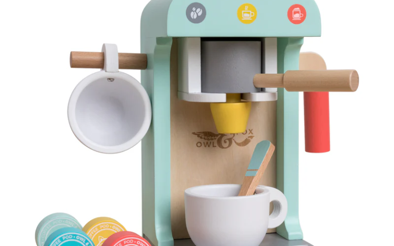 A wooden toy coffee machine in pastel colors with a cup, spoon, and wooden coffee pods. The machine has a handle, a cup holder on the side, and coffee-themed icons. Three coffee pods are on the base, each with animal faces and different colors.