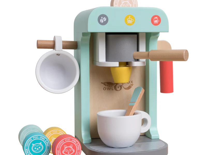 A wooden toy coffee machine in pastel colors with a cup, spoon, and wooden coffee pods. The machine has a handle, a cup holder on the side, and coffee-themed icons. Three coffee pods are on the base, each with animal faces and different colors.
