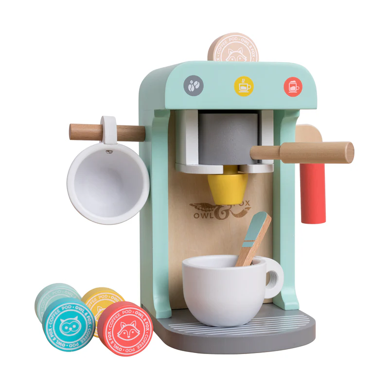 A wooden toy coffee machine in pastel colors with a cup, spoon, and wooden coffee pods. The machine has a handle, a cup holder on the side, and coffee-themed icons. Three coffee pods are on the base, each with animal faces and different colors.