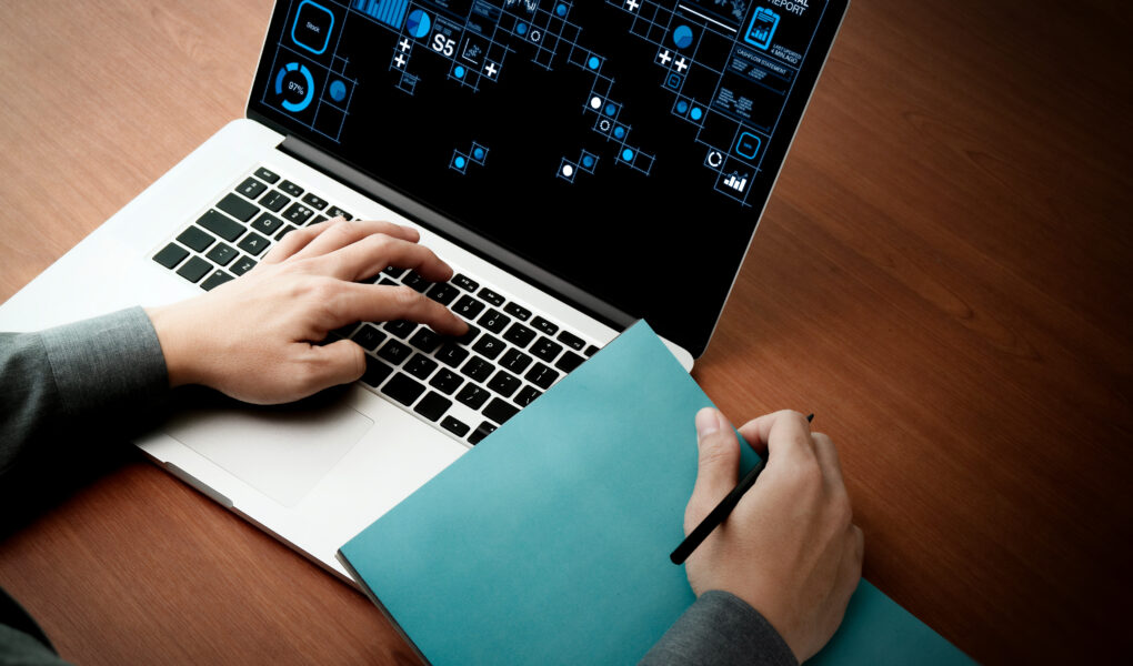 A person using a laptop with data analytics graphics on the screen, holding a pen and a blue folder on a wooden desk. One hand is typing on the keyboard while the other holds the pen, ready to write or take notes.