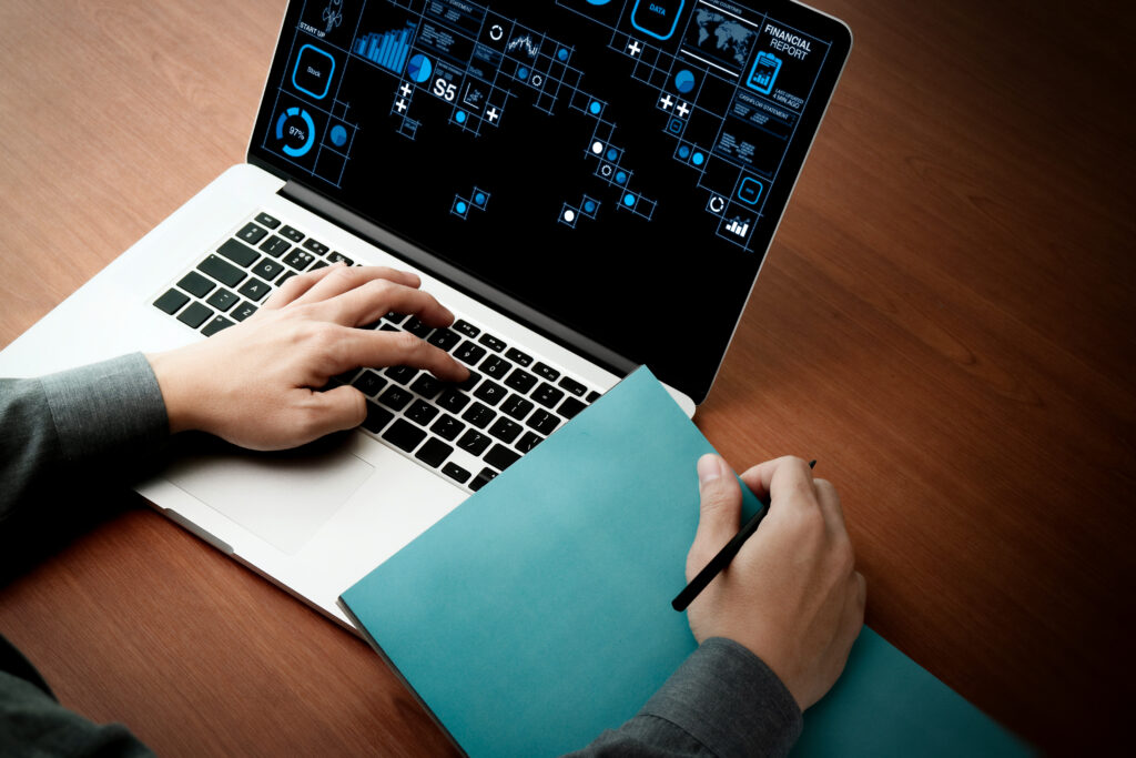 A person using a laptop with data analytics graphics on the screen, holding a pen and a blue folder on a wooden desk. One hand is typing on the keyboard while the other holds the pen, ready to write or take notes.