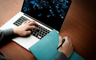 A person using a laptop with data analytics graphics on the screen, holding a pen and a blue folder on a wooden desk. One hand is typing on the keyboard while the other holds the pen, ready to write or take notes.