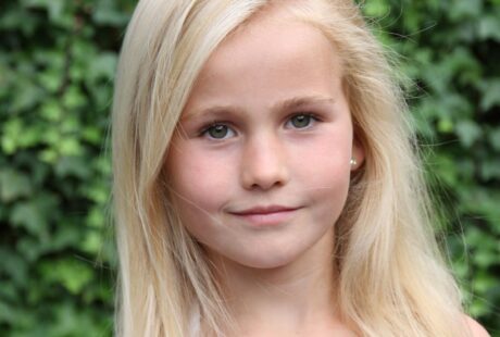 A young girl with long, straight blonde hair and blue eyes stands outdoors in front of a leafy green background. She is wearing a white top and has a calm, neutral expression on her face. The lighting is natural, highlighting her clear complexion.