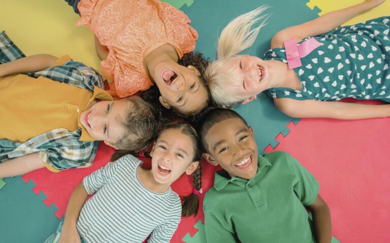 Five children lie on colorful foam mats in a circle with their heads together, smiling and laughing as they look up at the camera. The kids wear bright, casual clothes and appear joyful and playful, creating a cheerful and inclusive atmosphere.