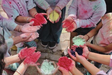 A group of people stand in a circle, holding out their hands filled with colorful powder in red, pink, green, yellow, and purple, ready to celebrate Holi, the festival of colors. Their clothes and arms are stained with powder, showing joyful participation.
