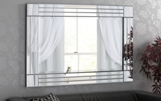 A decorative wall mirror reflects a window with sheer white curtains and part of a potted plant. The mirror hangs above a black leather sofa with a patterned cushion. The wall is covered in light grey wallpaper with a subtle floral design.