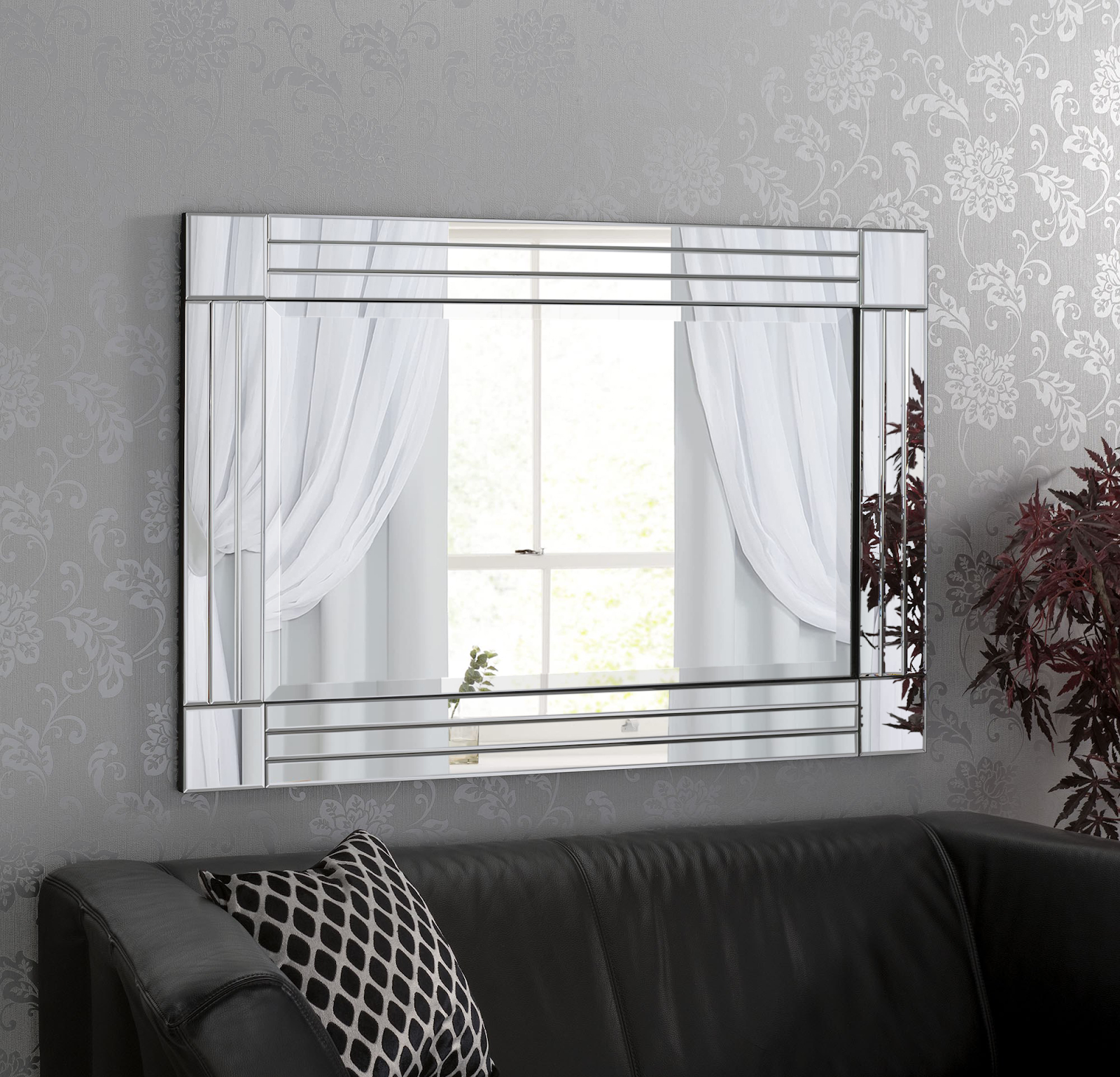 A decorative wall mirror reflects a window with sheer white curtains and part of a potted plant. The mirror hangs above a black leather sofa with a patterned cushion. The wall is covered in light grey wallpaper with a subtle floral design.