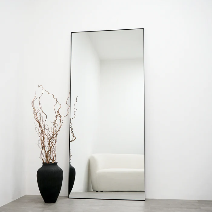 A large rectangular mirror with a thin black frame leans against a white wall. To the left of the mirror is a tall black vase holding dried curly branches. The mirror reflects a minimalist white sofa and the empty, bright room.