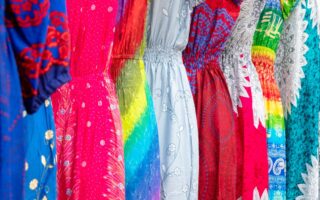 A row of colorful dresses hangs side by side. Each dress features a unique pattern, including floral, tie-dye, feathers, and geometric shapes in vibrant shades of blue, pink, red, green, yellow, and white. The fabrics are light and gathered at the waist.