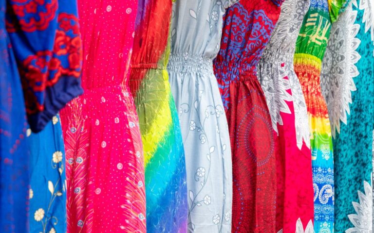 A row of colorful dresses hangs side by side. Each dress features a unique pattern, including floral, tie-dye, feathers, and geometric shapes in vibrant shades of blue, pink, red, green, yellow, and white. The fabrics are light and gathered at the waist.