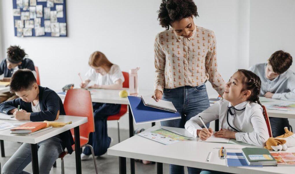 A teacher stands beside a seated student, smiling and holding an open folder. The student, a girl, smiles up while writing. Other students sit at desks, focused on their work. The classroom has red chairs, white desks, and a bulletin board in the background.