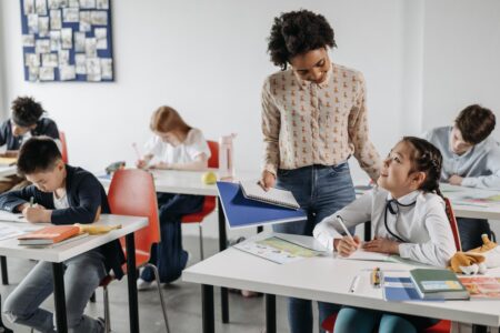 A teacher stands beside a seated student, smiling and holding an open folder. The student, a girl, smiles up while writing. Other students sit at desks, focused on their work. The classroom has red chairs, white desks, and a bulletin board in the background.
