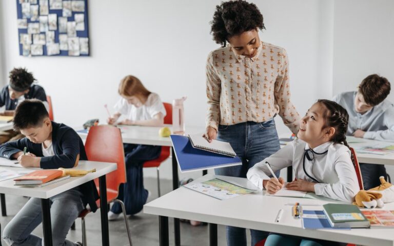A teacher stands beside a seated student, smiling and holding an open folder. The student, a girl, smiles up while writing. Other students sit at desks, focused on their work. The classroom has red chairs, white desks, and a bulletin board in the background.