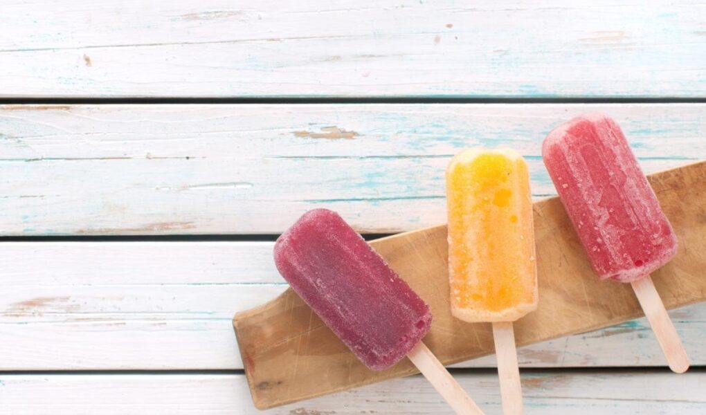 Three colorful popsicles—purple, yellow, and red—rest on a wooden board atop a white rustic wooden surface. The popsicles appear frosty and refreshing, with their sticks facing forward, creating a vibrant and summery scene.