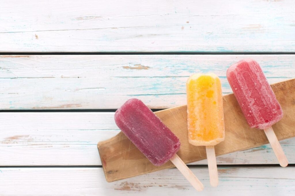 Three colorful popsicles—purple, yellow, and red—rest on a wooden board atop a white rustic wooden surface. The popsicles appear frosty and refreshing, with their sticks facing forward, creating a vibrant and summery scene.