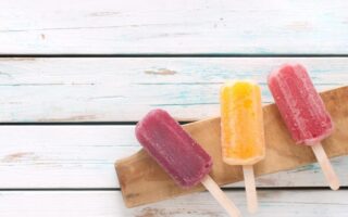 Three colorful popsicles—purple, yellow, and red—rest on a wooden board atop a white rustic wooden surface. The popsicles appear frosty and refreshing, with their sticks facing forward, creating a vibrant and summery scene.