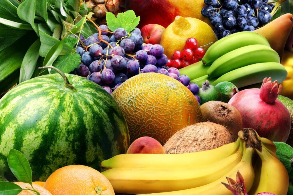 A colorful assortment of fresh fruits including watermelon, bananas, green and purple grapes, coconut, pomegranate, orange, kiwi, melon, lemon, pear, apple, peaches, blueberries, and cherries, arranged closely together.