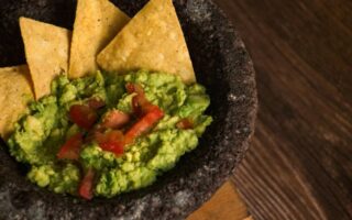 A stone bowl filled with chunky guacamole, topped with diced tomatoes, sits on a wooden surface. Three yellow tortilla chips are dipped into the guacamole, and the lighting highlights the fresh texture and vibrant colors of the ingredients.