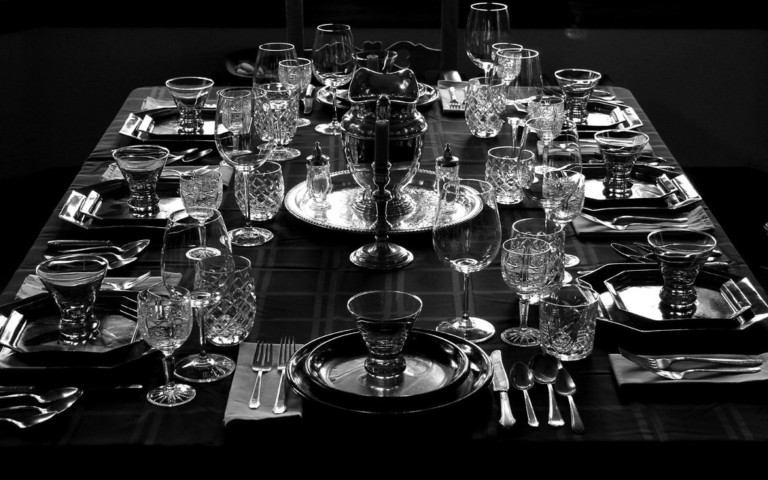 A black-and-white photo of an elegant dining table set for eight, with ornate glassware, plates, cutlery, folded napkins, and a reflective centerpiece. The arrangement is symmetrical and formal, creating a sense of sophistication and anticipation.