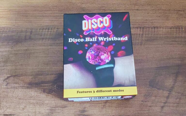 A rectangular box labelled "DISCO Disco Ball Wristband" sits on a wooden surface. The box shows a photo of a lit, pink mini disco ball on a wristband, emitting colourful light spots. Text below reads, "Features 3 different modes.