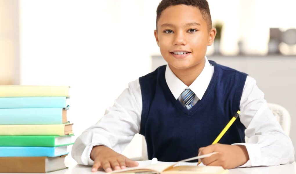 A smiling boy wearing a white shirt, blue waistcoat, and striped tie sits at a desk, holding a pencil over an open exercise book. A stack of colourful books is beside him. The background is softly blurred, suggesting an indoor, school-like environment.