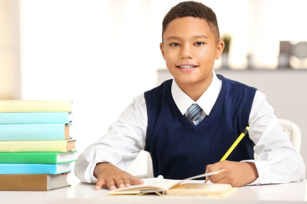 A young boy wearing a white shirt, blue waistcoat, and striped tie sits at a desk, smiling, and writes in an open exercise book with a yellow pencil. A stack of colourful books is on the desk beside him. The background is bright and blurred.
