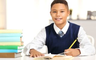A young boy wearing a white shirt, blue waistcoat, and striped tie sits at a desk, smiling, and writes in an open exercise book with a yellow pencil. A stack of colourful books is on the desk beside him. The background is bright and blurred.