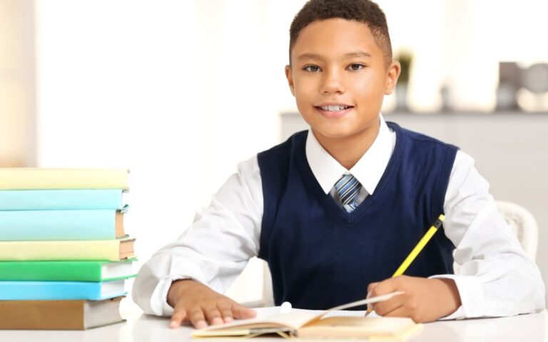 A young boy wearing a white shirt, blue waistcoat, and striped tie sits at a desk, smiling, and writes in an open exercise book with a yellow pencil. A stack of colourful books is on the desk beside him. The background is bright and blurred.