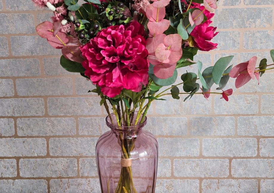 A bouquet of artificial flowers in a purple glass vase sits on a wooden surface against a grey brick wall. The arrangement features deep pink peonies, soft pink fluffy blossoms, and stems of eucalyptus with round, dusty green and reddish leaves.